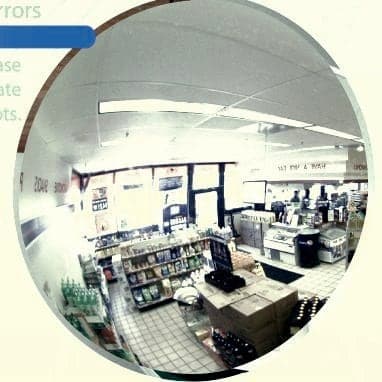 Round convex mirror mounted high on a convenience store wall showing the register, coolers, and aisle ends in one view