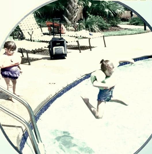 Convex mirror view of an outdoor pool deck showing a child jumping into the water and another child at the edge